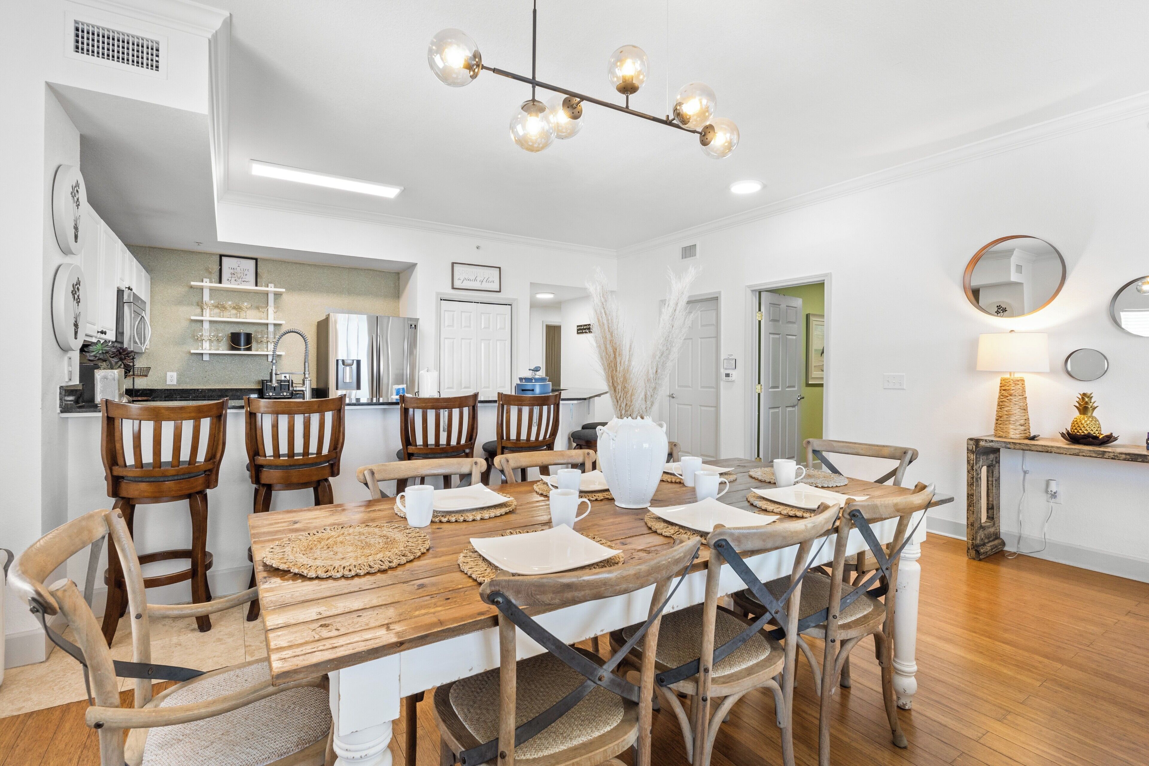 A dining area with a wooden table and chairs, a chandelier, and a kitchen in the background.