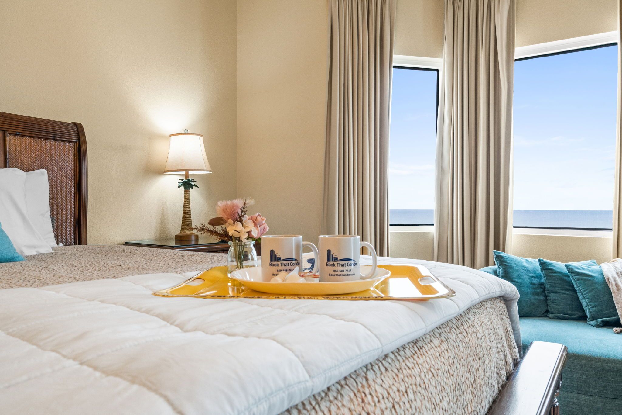 A hotel room with a bed, a bedside table with a lamp, a tray with two mugs, and a view of the sea through large windows.