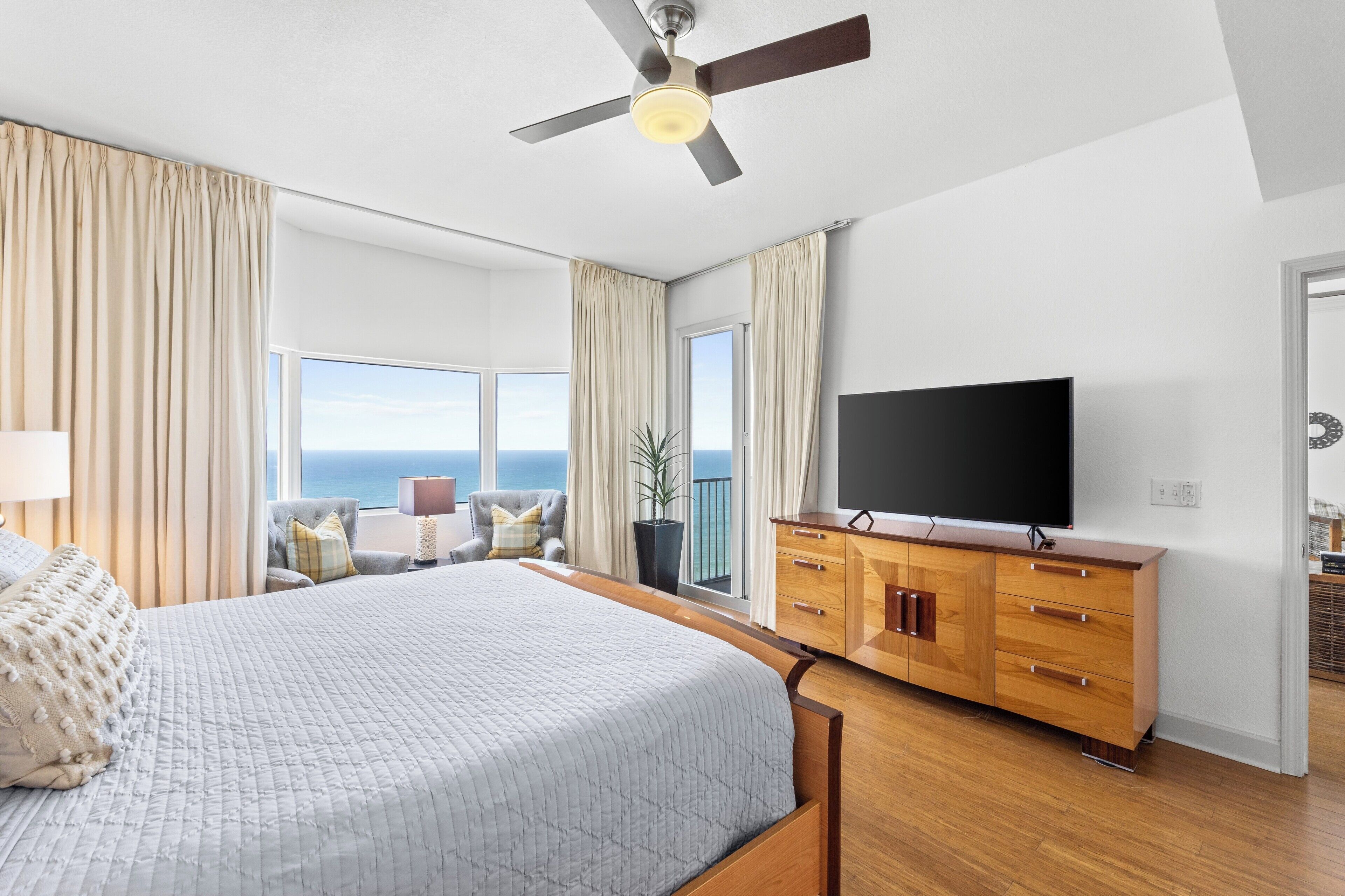 A spacious bedroom with a large bed, a flat-screen TV, wooden furniture, and a view of the ocean.