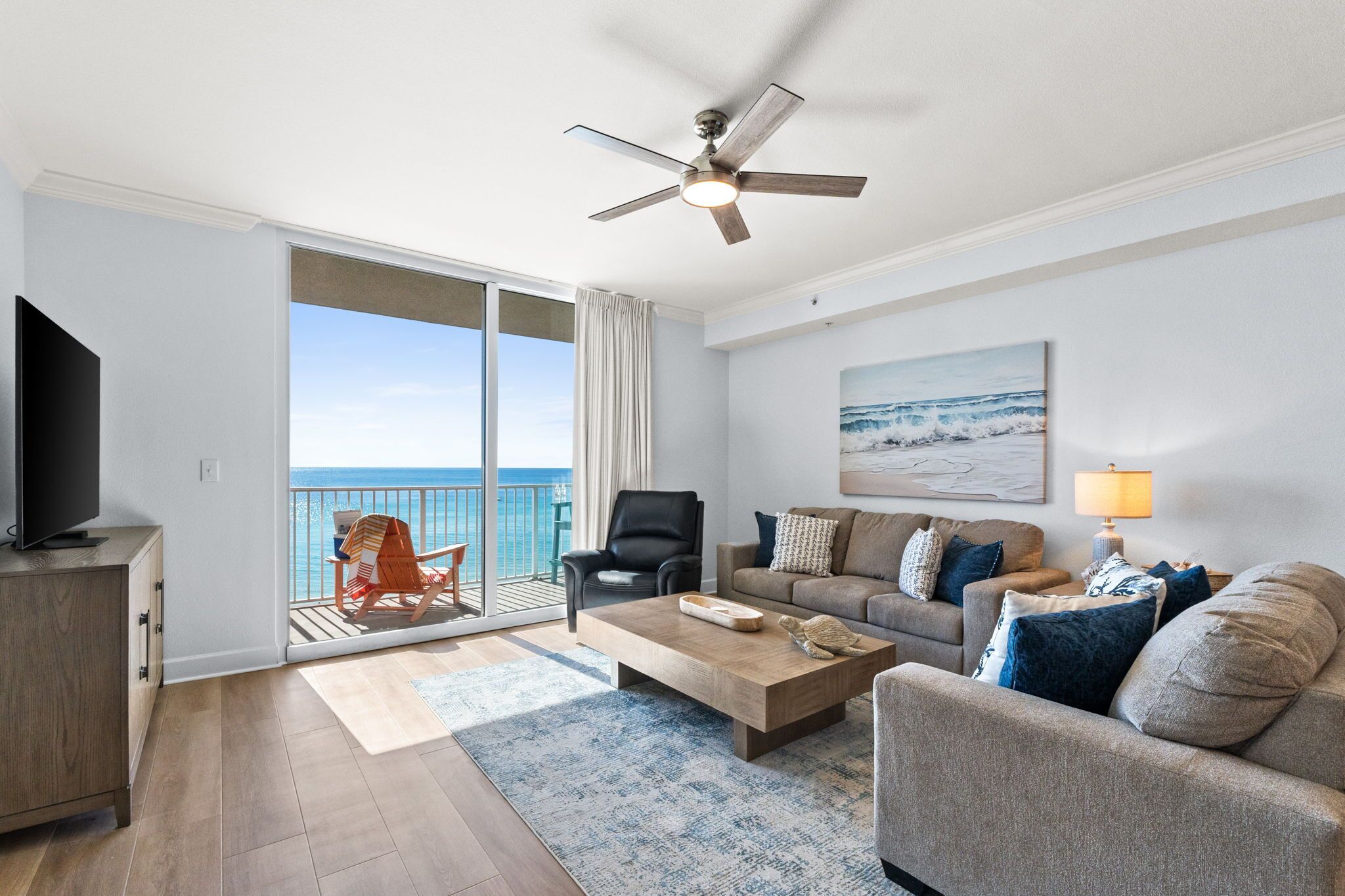 A spacious living room with a large TV, a ceiling fan, a sofa, a coffee table, and a view of the ocean through a sliding glass door.