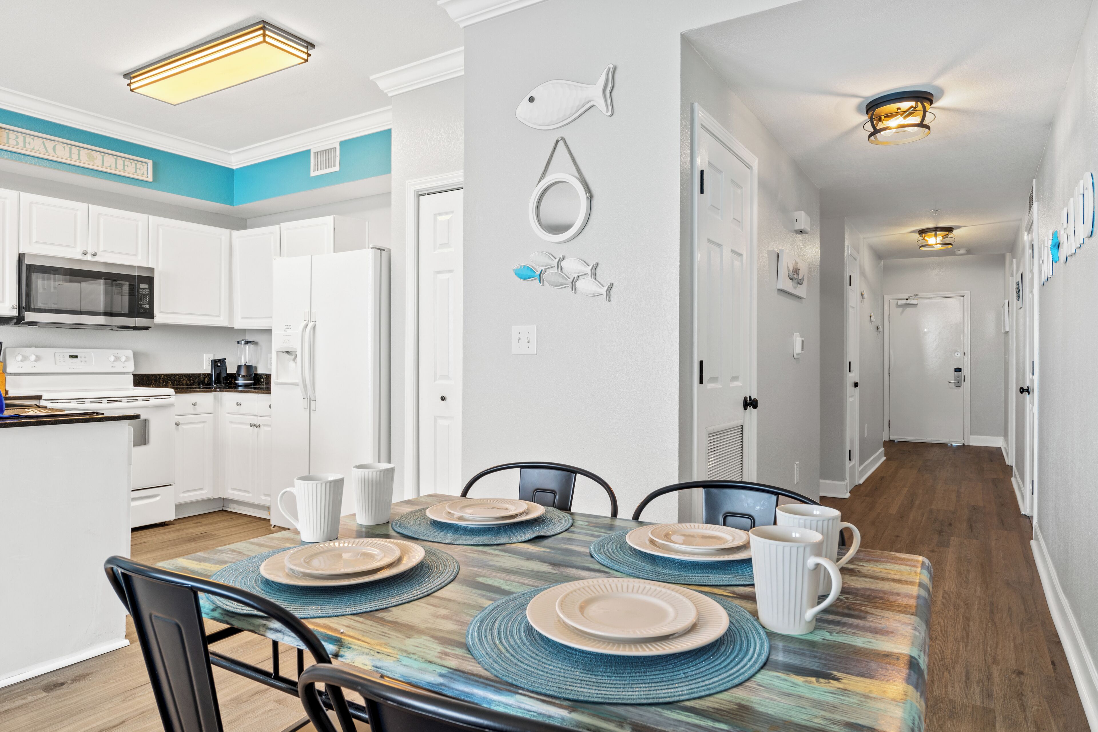 A modern kitchen with white cabinets, a microwave, and a refrigerator. There is a dining table set with plates and cups.