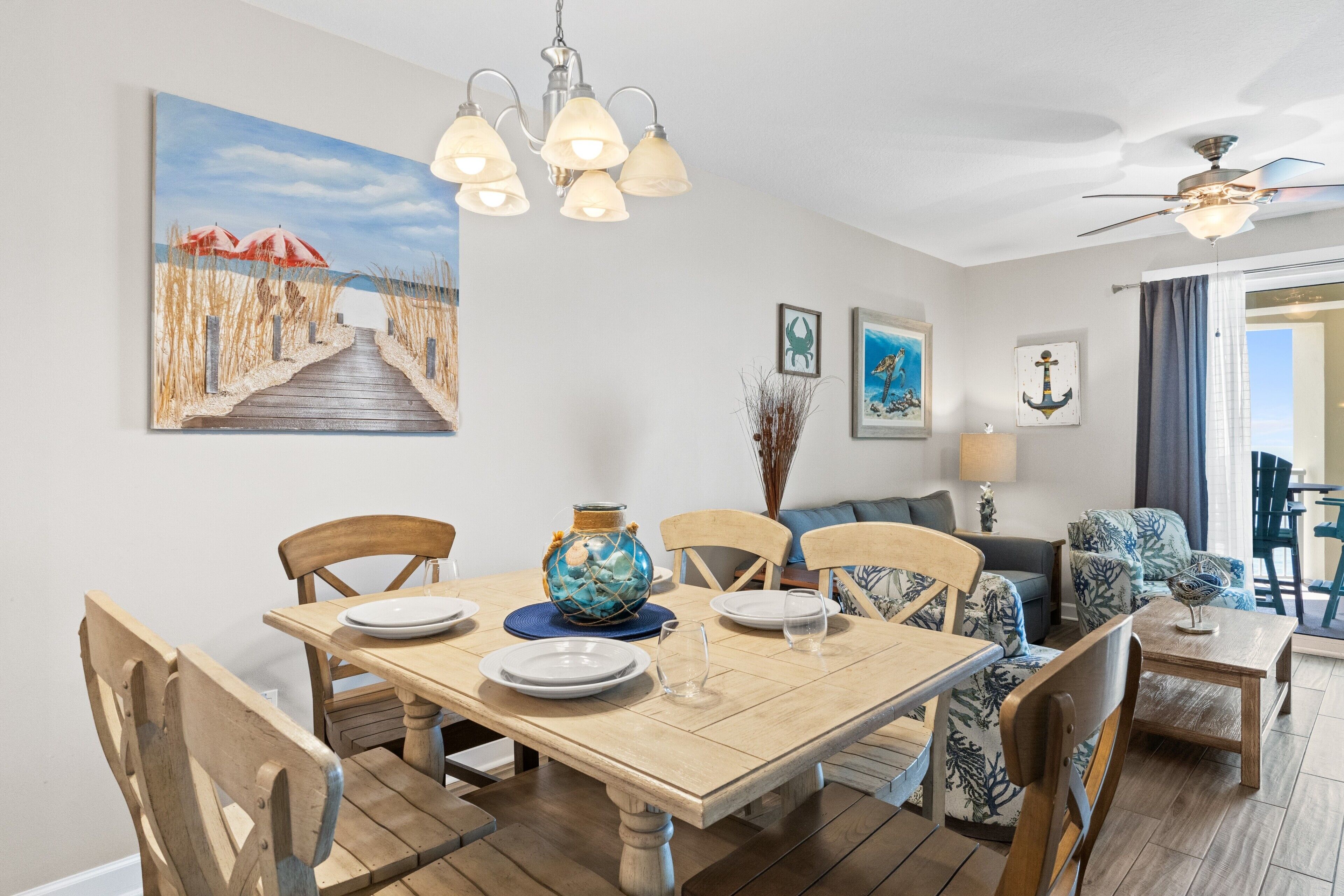 A dining area with a wooden table set for four, surrounded by wooden chairs. A painting of a beach scene is on the wall, and there is a ceiling fan above the table.