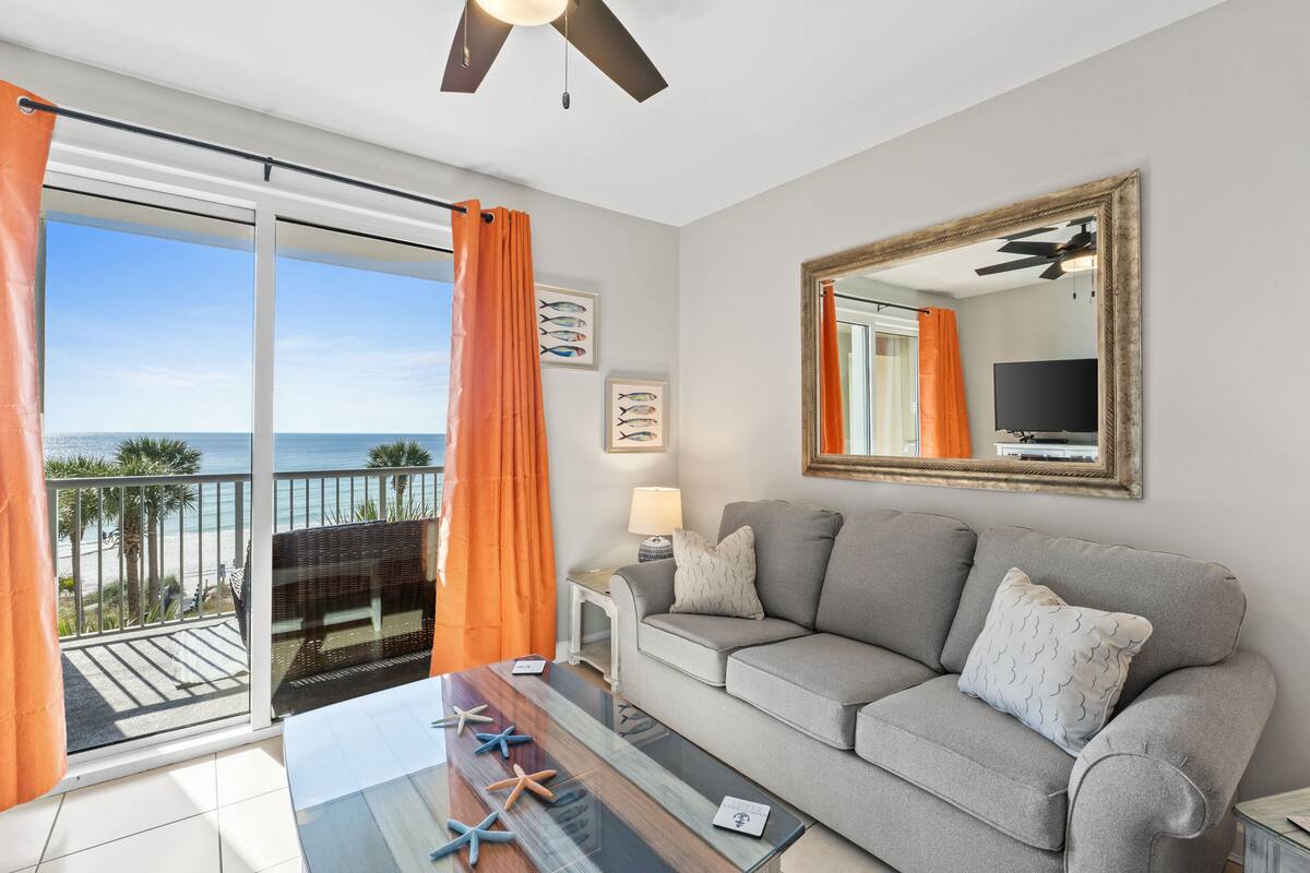 Un balcon avec vue sur l’océan, un canapé gris, une table basse en verre ornée d’étoiles de mer et un ventilateur de plafond.