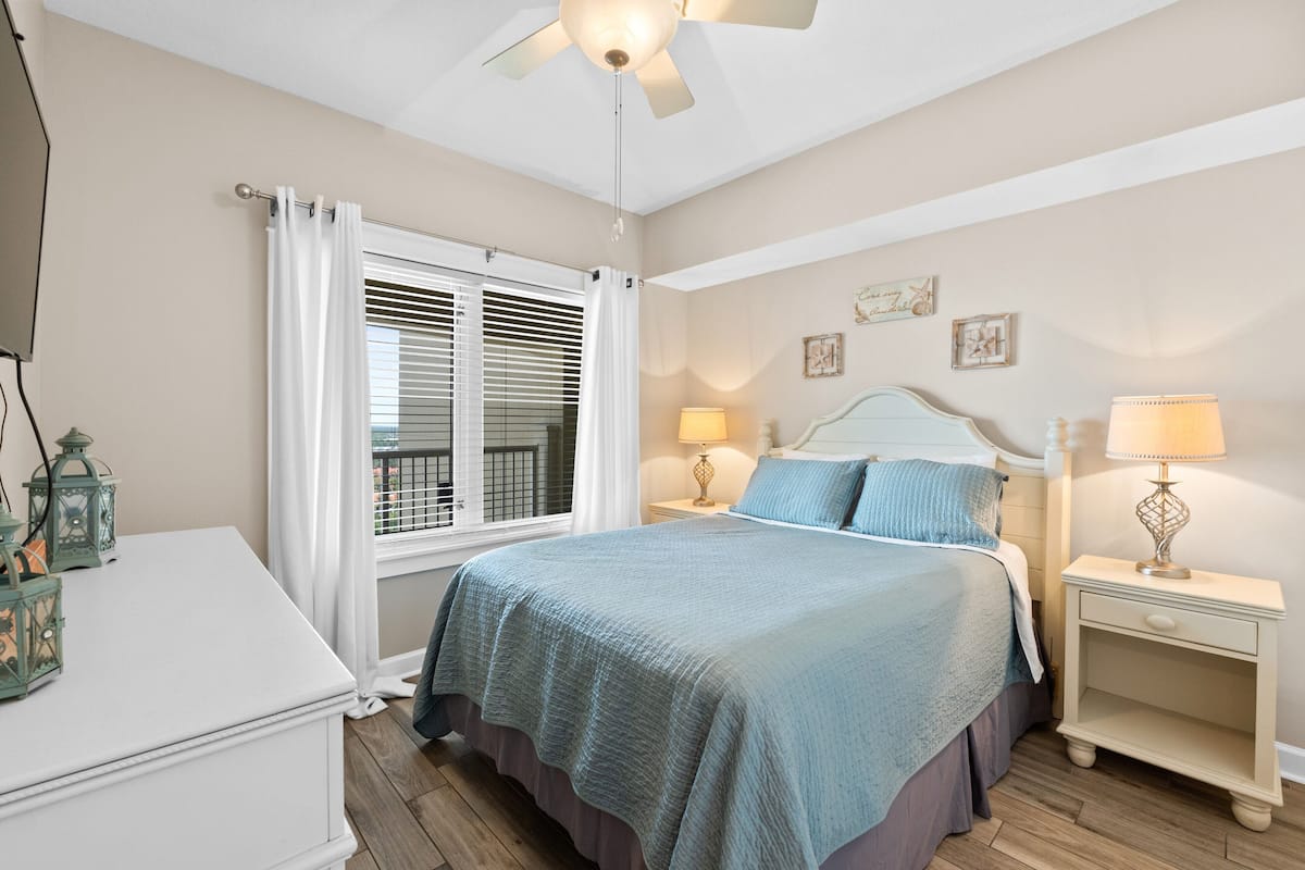 Une chambre à coucher comprenant un lit, des tables de chevet, une commode et une fenêtre avec des stores.