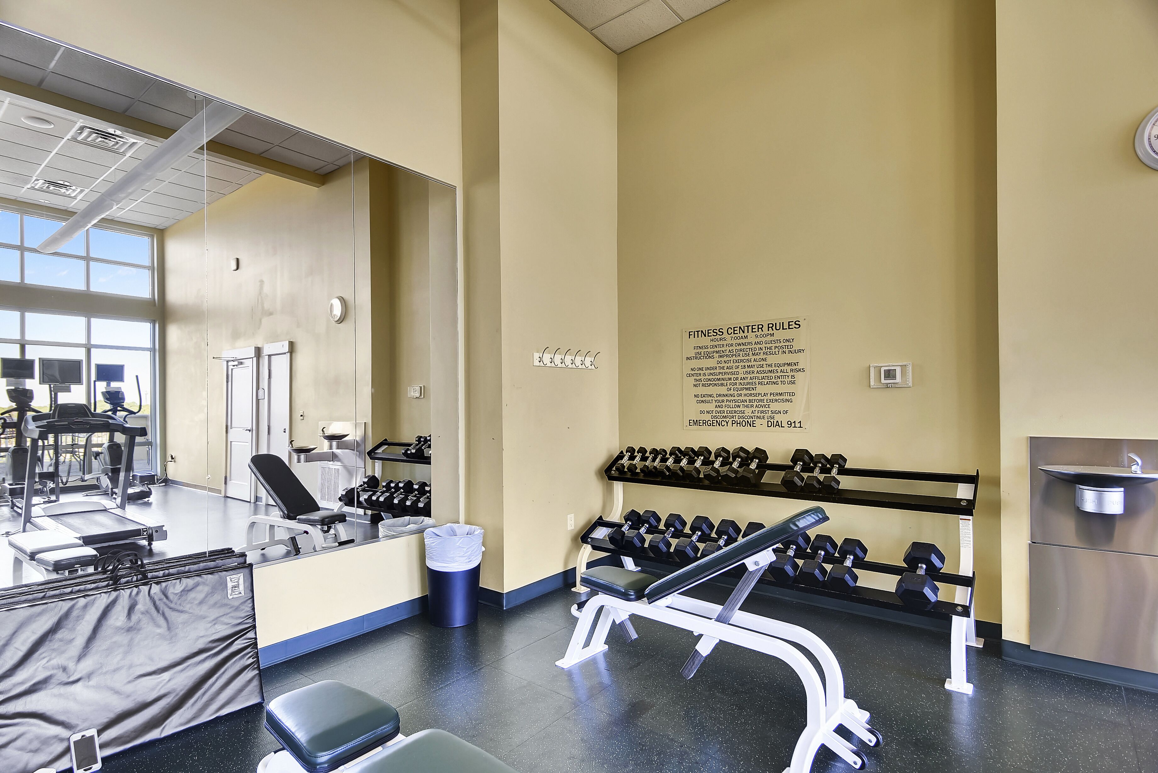 A fitness center with exercise equipment, a wall-mounted sign with rules, and a trash can.