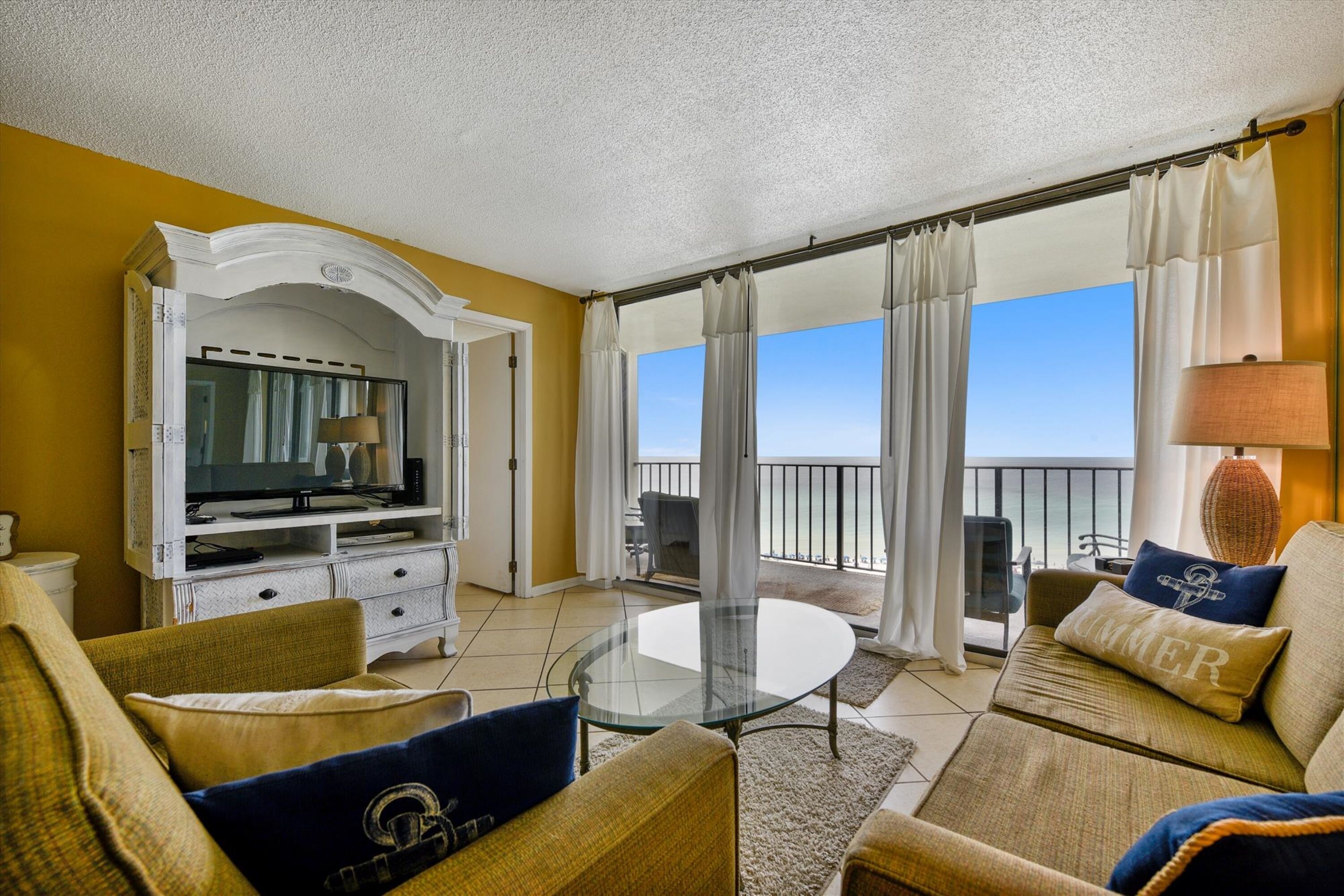A living room with a glass-top coffee table, sofas, a television, and a balcony with curtains.