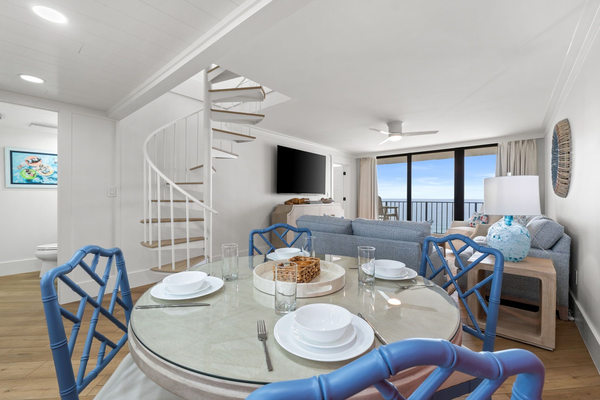 Un salon moderne avec un escalier en colimaçon, un coin repas avec une table ronde et des chaises, un ensemble de canapés et un téléviseur fixé au mur.