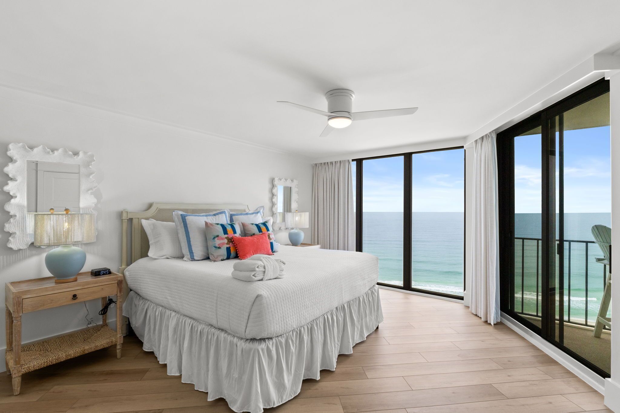 A bedroom with a large bed, a ceiling fan, a nightstand, and a view of the ocean.