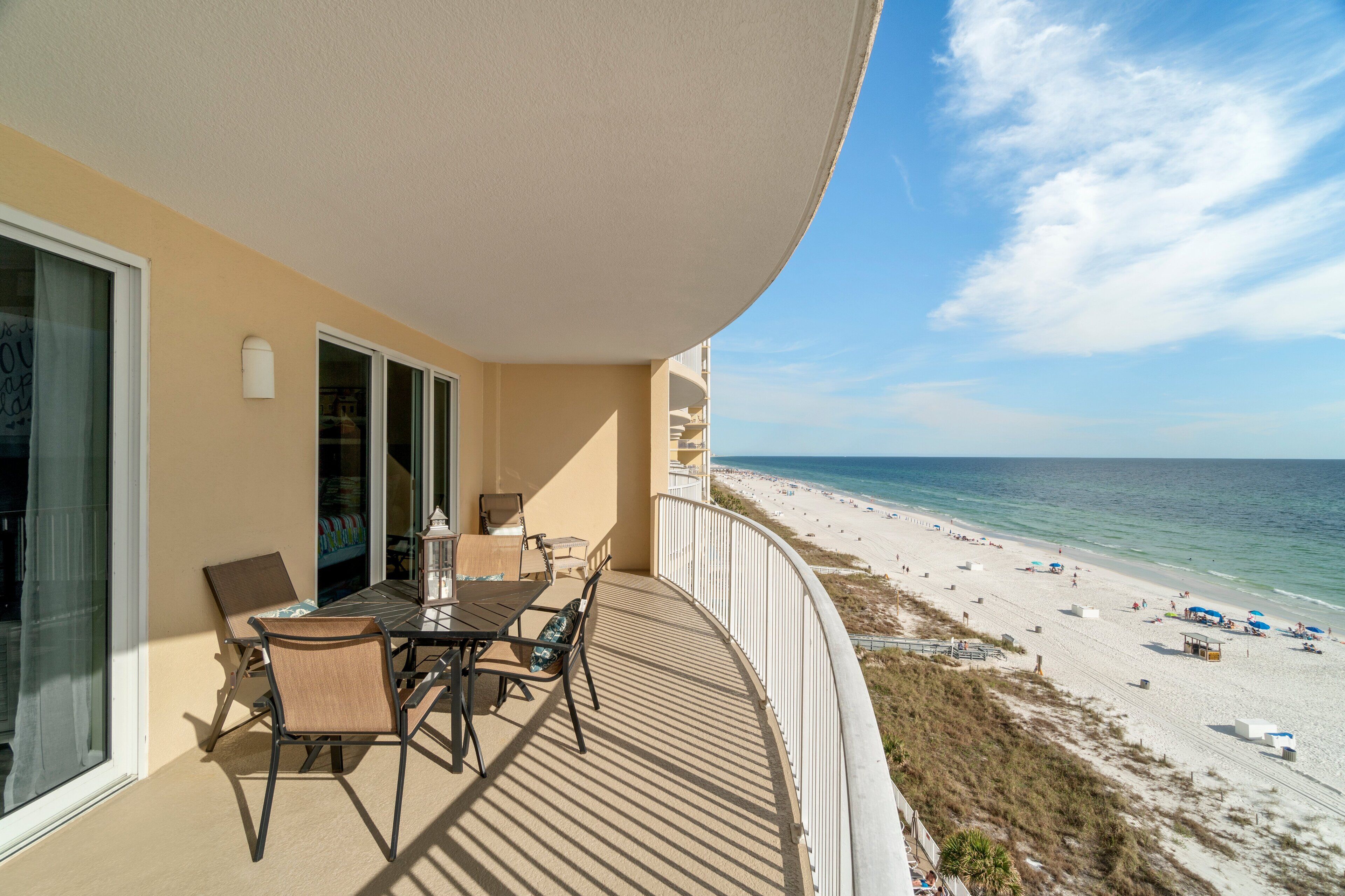 Een balkon met een tafel en stoelen, met uitzicht op een strand met parasols en mensen.