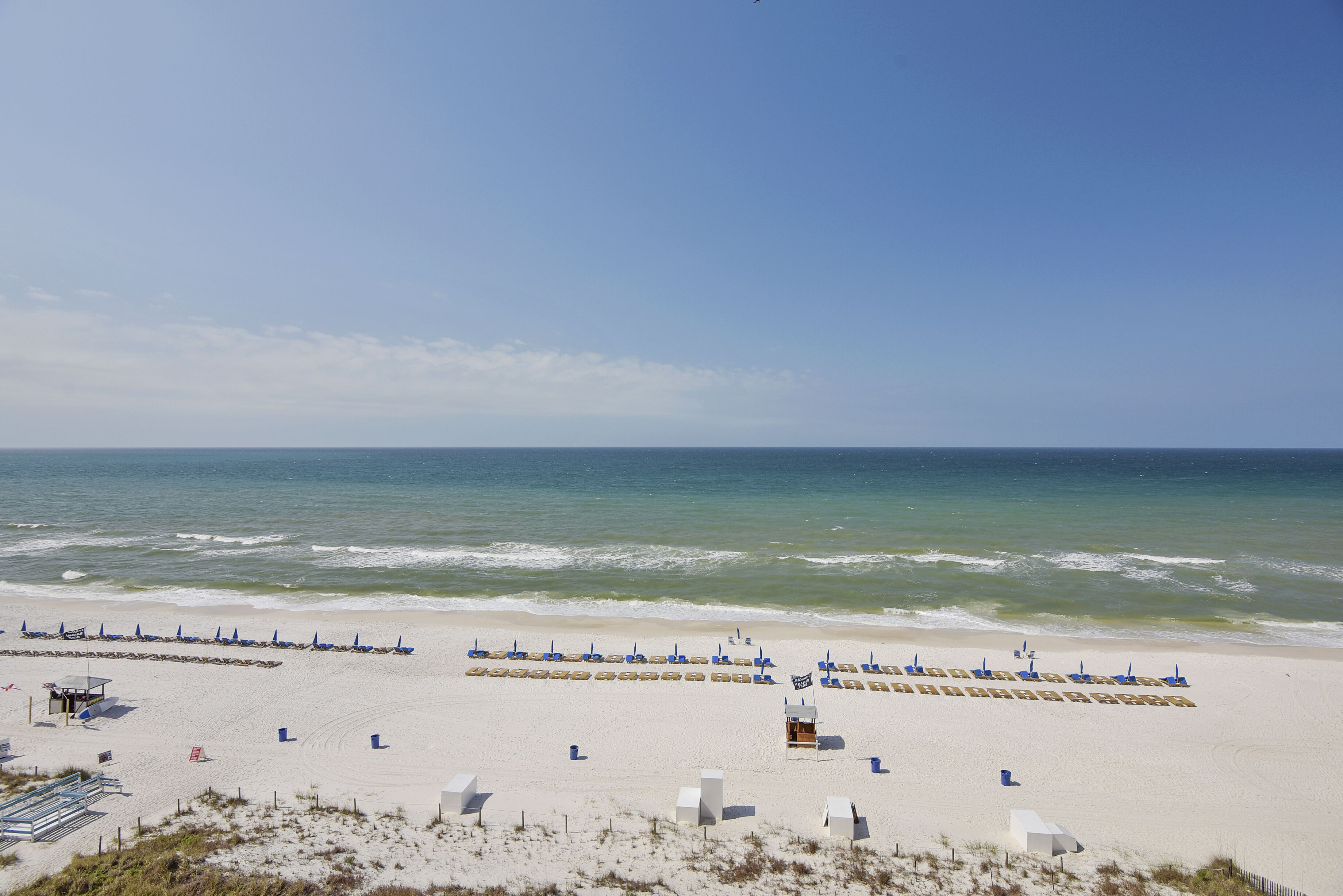Une plage bordée de rangées de chaises de plage et de parasols bleus, avec une tour de surveillance et une zone de plage sablonneuse.