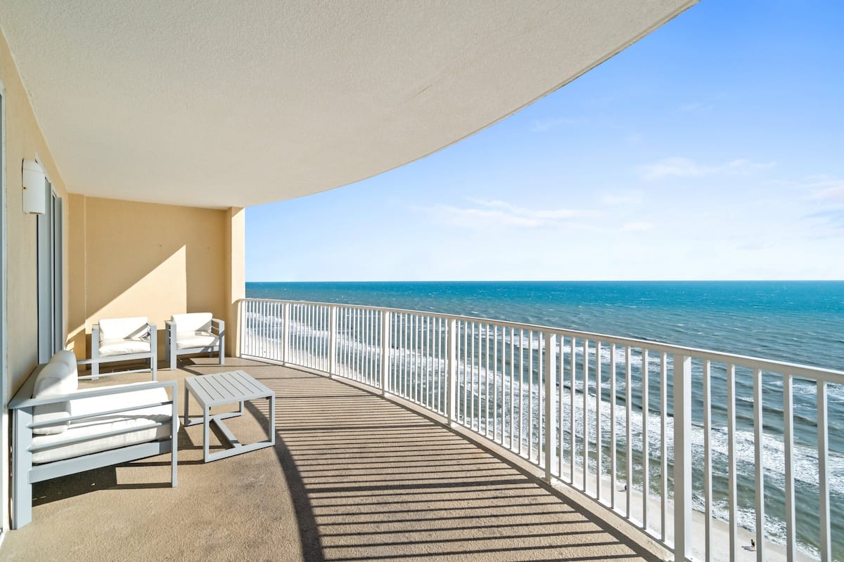 Un balcón con sillas y una mesa blancos, con vista a una playa y una barandilla.