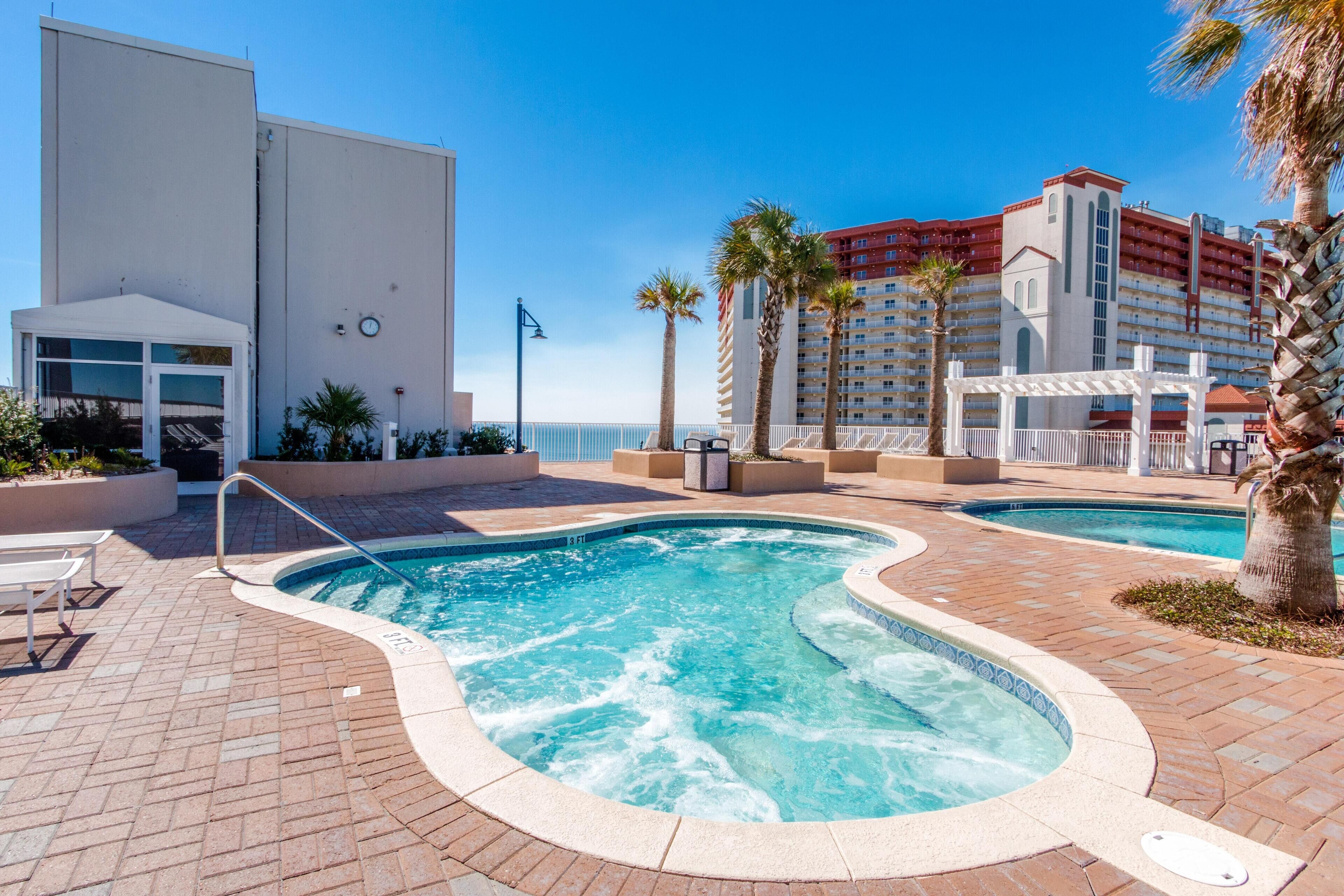 A pool area with two hot tubs, a white building, and a palm tree.