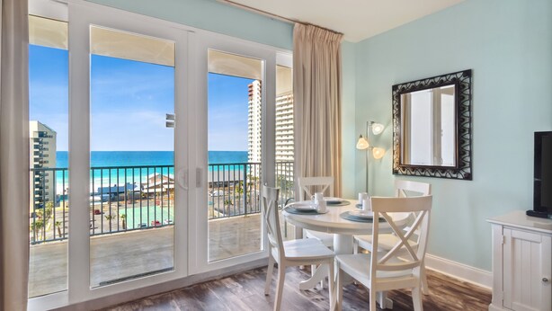 A room with a balcony overlooking a beach and ocean, a dining table with chairs, a TV, and a cabinet.