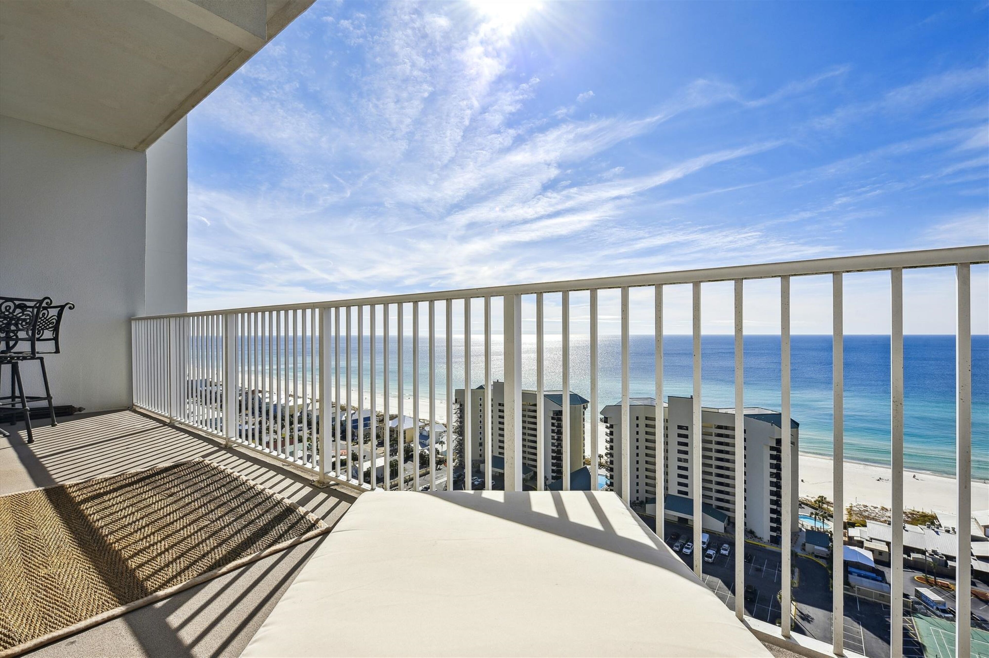 A balcony with a view of the beach and ocean, featuring a white railing and a woven mat.