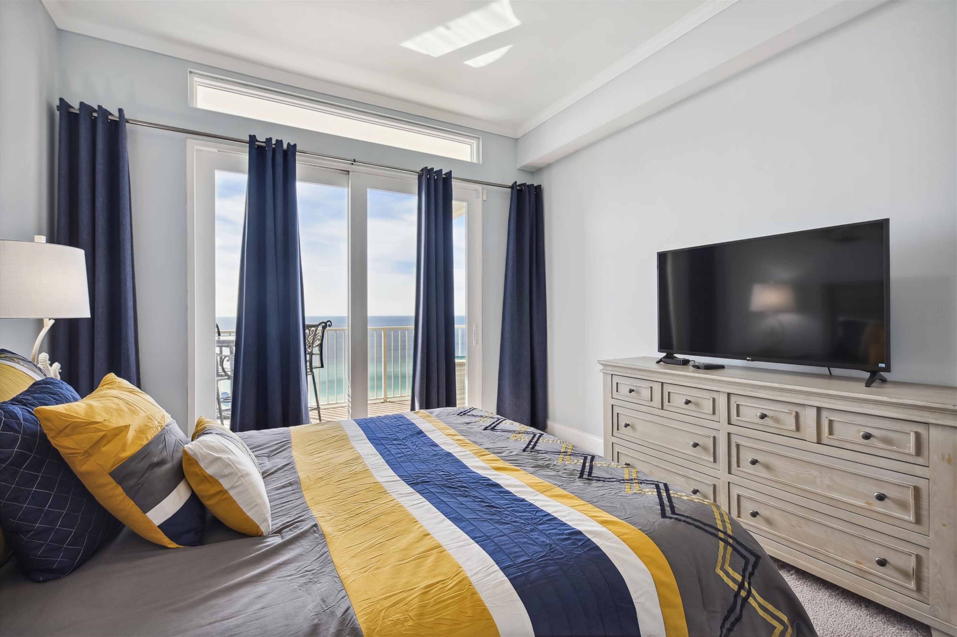 A hotel room with a bed, a TV, a dresser, and a balcony with curtains.