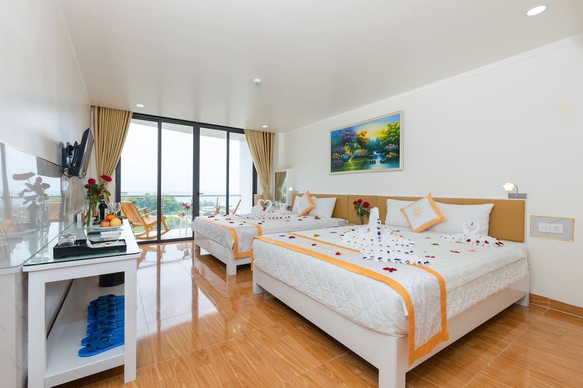 A hotel room with two beds, a glass table with a vase of flowers, a painting on the wall, and a view of the outdoors.