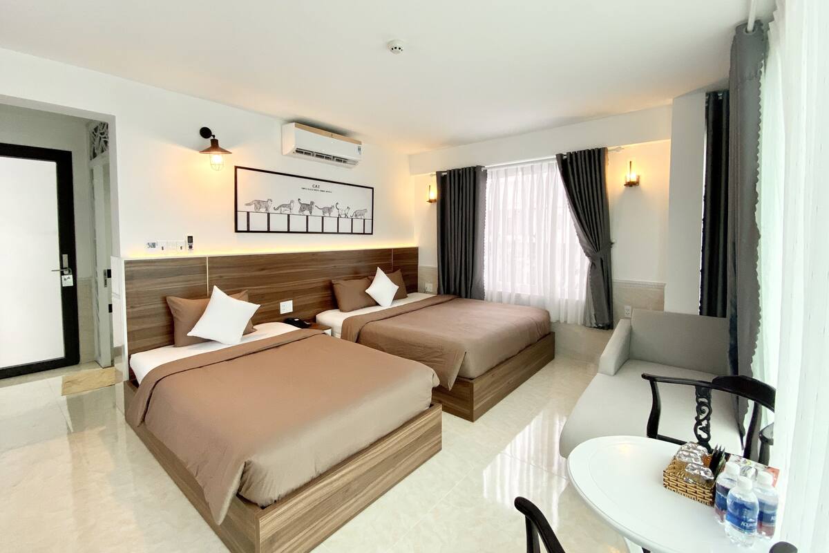 A hotel room with two beds, a small table, a chair, a desk, and a window with curtains.