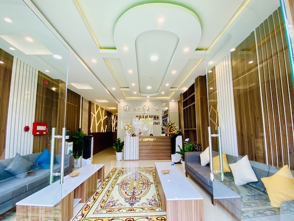 A hotel reception area with a modern design, featuring a reception desk, sofas, a patterned rug, and a decorative wall.