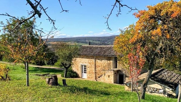 Une maison en pierre avec un toit de tuiles, entourée d’un champ verdoyant et d’arbres aux feuilles d’automne.
