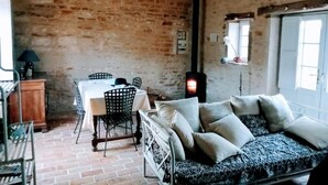 Une pièce rustique avec un mur en briques, un lit recouvert d’une literie à motifs, une table avec des chaises et un poêle à bois.