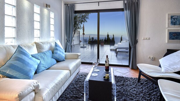 A modern living room with a white sofa, a glass coffee table, and a view of the outdoors through large sliding doors. - Finca Stemann - Casa Pascal (Guia de Isora)
