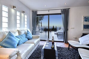 Ein modernes Wohnzimmer mit einem weißen Sofa, einem Glastisch und Blick ins Freie durch große Schiebetüren.