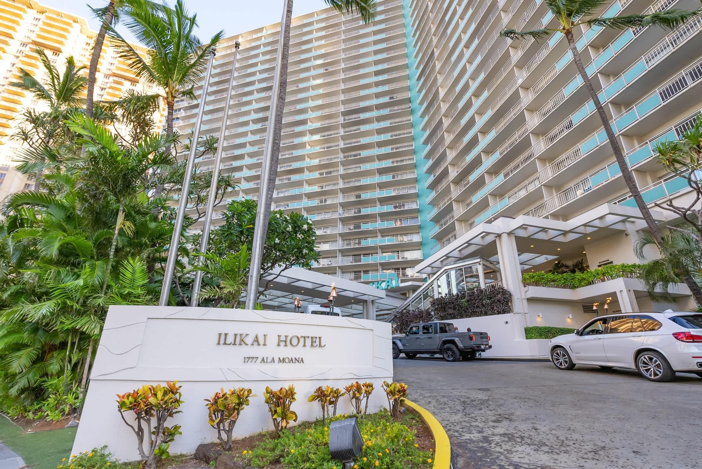 The Ilikai Hotel entrance with a sign, surrounded by palm trees and a parking area with cars.