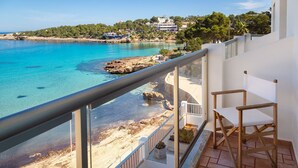 A balcony with a view of a turquoise sea, rocky shoreline, and buildings in the background. - Hostal La Cigüeña (Sant Joan de Labritja)