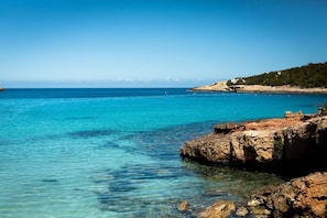 Kustlandschap met helder turquoise water, een rotsachtige kustlijn en een heuvel in de verte met schaarse begroeiing.