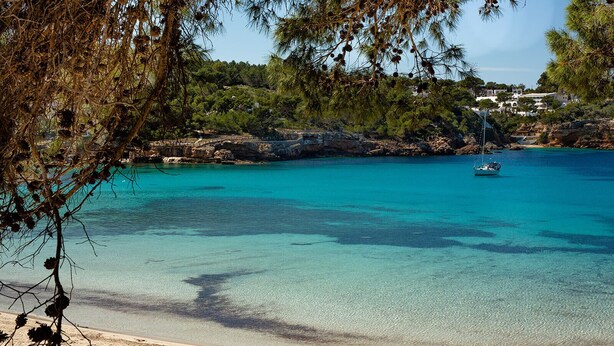 A beach with clear turquoise water, a boat, and rocky cliffs with buildings in the background. - Hostal La Cigüeña (Sant Joan de Labritja)