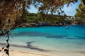 Ein Strand mit klarem türkisblauem Wasser, einem Boot und felsigen Klippen mit Gebäuden im Hintergrund.