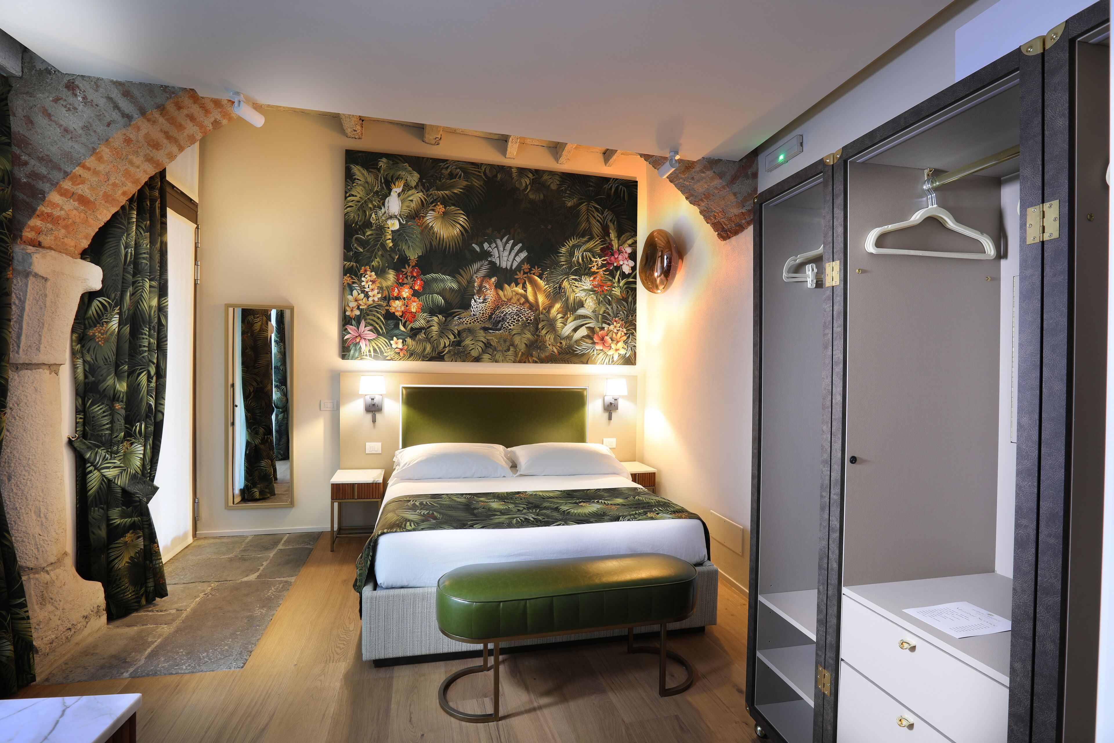 A hotel room with a bed, a bench, a wardrobe, a mirror, and a tropical-themed painting on the wall.