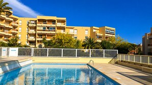 A swimming pool with a clear blue water surface, surrounded by a white fence. In the background, there are multi-story buildings with balconies and a clear blue sky. - l'Appartement Magnolia (Vallauris)