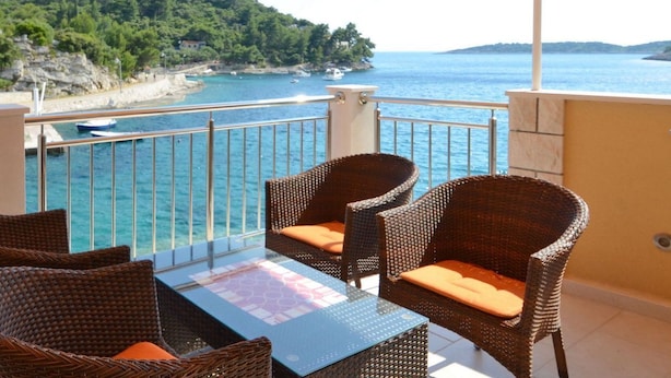 Un balcon agrémenté de fauteuils en osier et d’une table en verre, offrant une vue sur une zone côtière avec des bateaux et de la végétation.