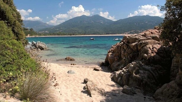 Una playa con aguas turquesas cristalinas, formaciones rocosas y montañas a lo lejos.
