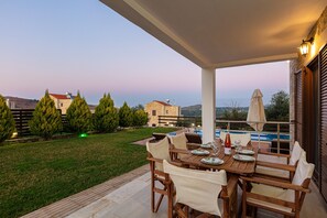 A covered outdoor dining area with a pool view, wooden furniture, and a stone building in the background.