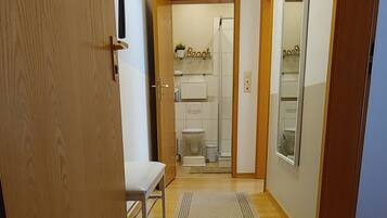 Un pasillo con felpudo, banco y vistas a un baño con inodoro y ducha.