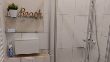 Une salle de bain avec une douche à parois de verre, des toilettes, un petit tabouret et une étagère sur laquelle se trouvent une plante et un panneau indiquant « Plage ».