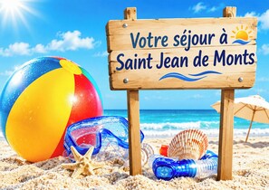 Eine Strandansicht mit einem Holzschild, auf dem 'Votre séjour à Saint Jean de Monts' steht, und einem bunten Strandball.