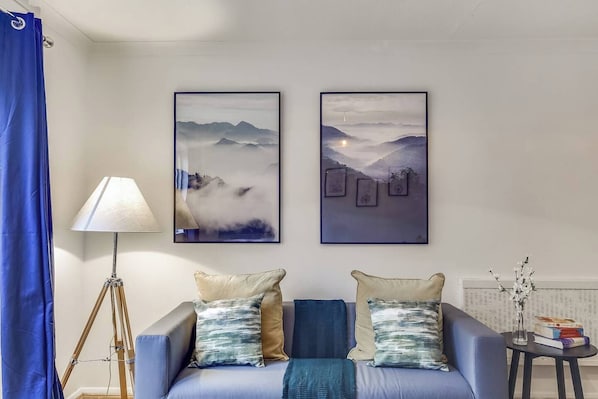 A living room with a sofa, two framed artworks depicting mountainous landscapes, a floor lamp, and a small table with books and a vase of flowers.