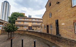 A courtyard with a brick building, a metal fence, and a tall residential building in the background. - Stylish 4BD Home with Garden Near Kennington (London)