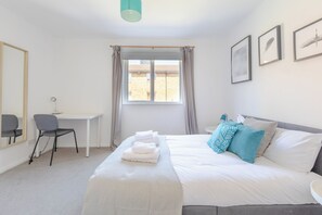 A hotel room with a bed, a desk, a chair, a window with curtains, and framed pictures on the wall. - Stylish 4BD Home with Garden Near Kennington (London)