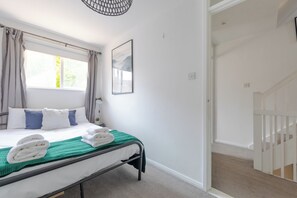 A bedroom with a bed, a window with curtains, a painting on the wall, and a staircase leading to another room.