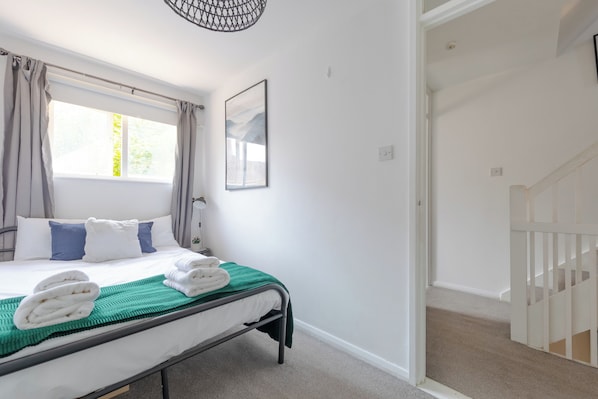 A bedroom with a bed, a window with curtains, a painting on the wall, and a staircase leading to another room.