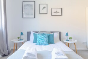 A neatly made bed with white linens, two pillows, and a pair of folded towels. There are two bedside tables with lamps and small potted plants. The room has a simple, clean design with a light-colored wall and a framed picture above the bed.
