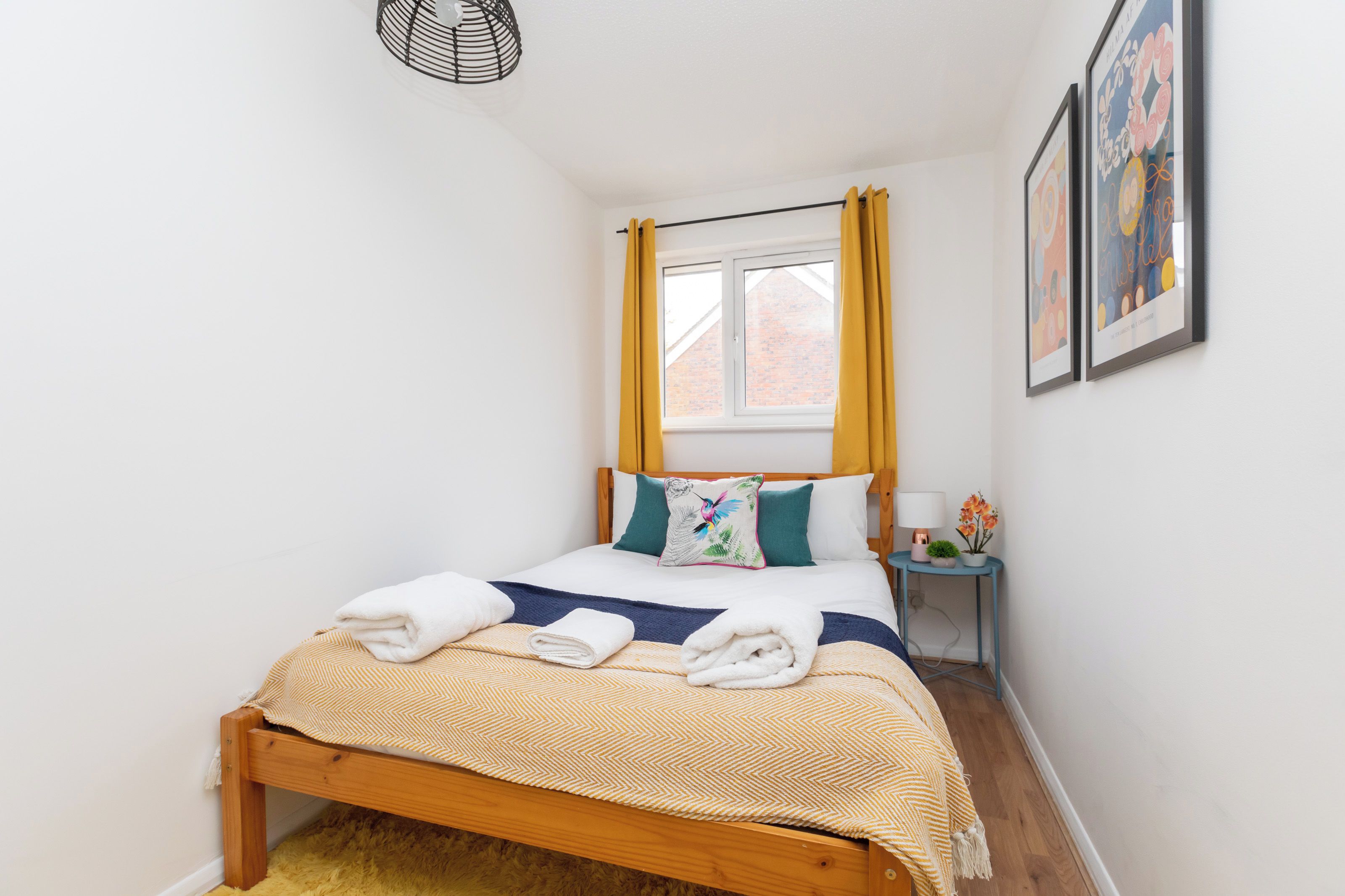 A neatly made bed with a wooden frame, a window with yellow curtains, a small bedside table with a lamp, and a wall with framed artwork.