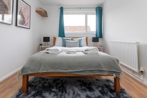 Un lit bien fait, recouvert d’une couverture grise, deux lampes de chevet, une fenêtre avec des rideaux de couleur turquoise et un radiateur.