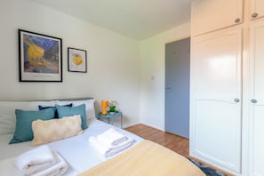 A hotel room with a bed, pillows, a nightstand with a lamp, a framed painting, and a wardrobe.