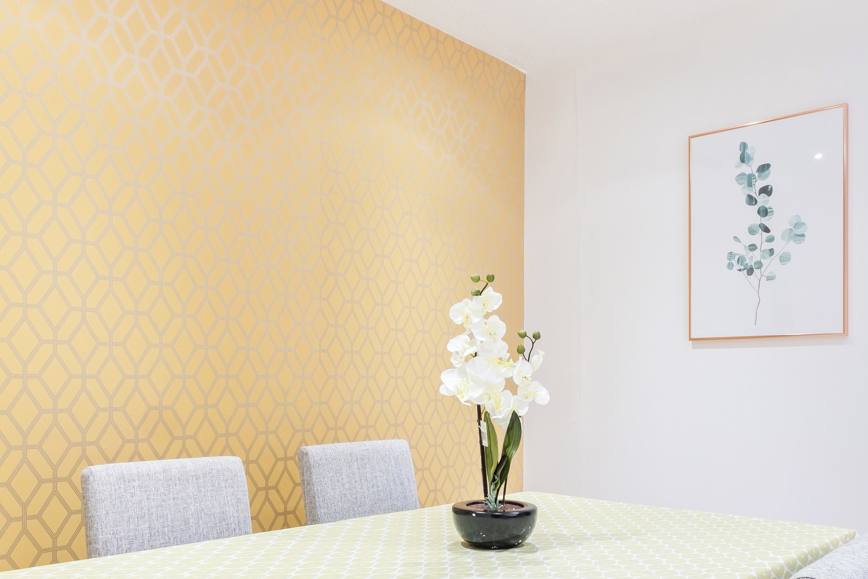A dining area with a table, chairs, a vase with white flowers, and a framed artwork on the wall.