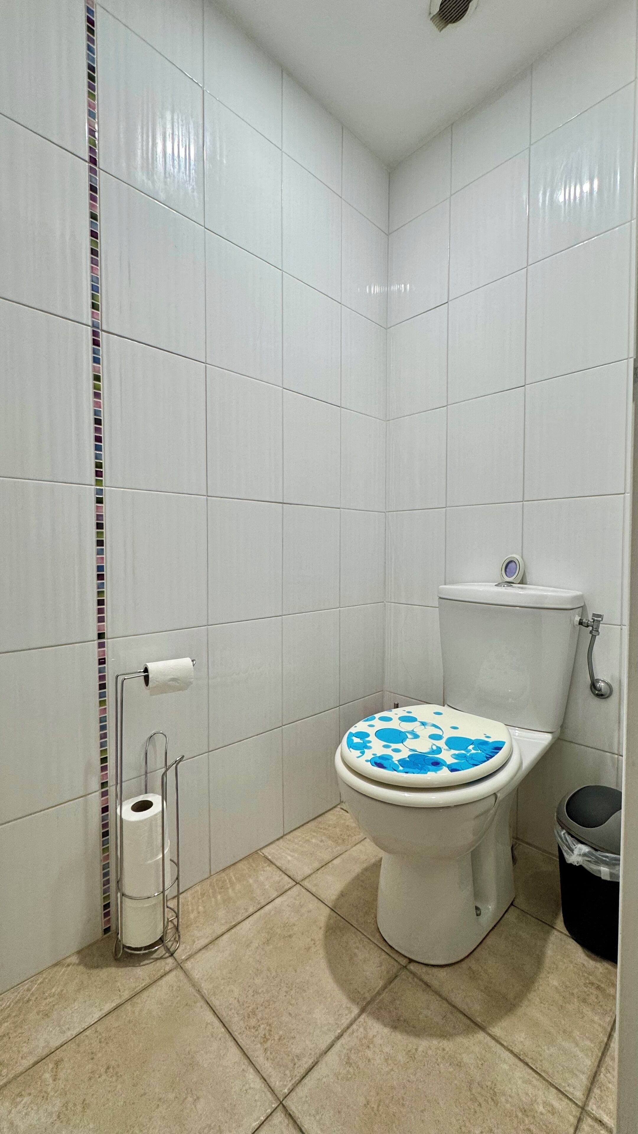 A bathroom with white tiled walls, a toilet with a blue patterned seat, a toilet paper holder, and a trash bin.